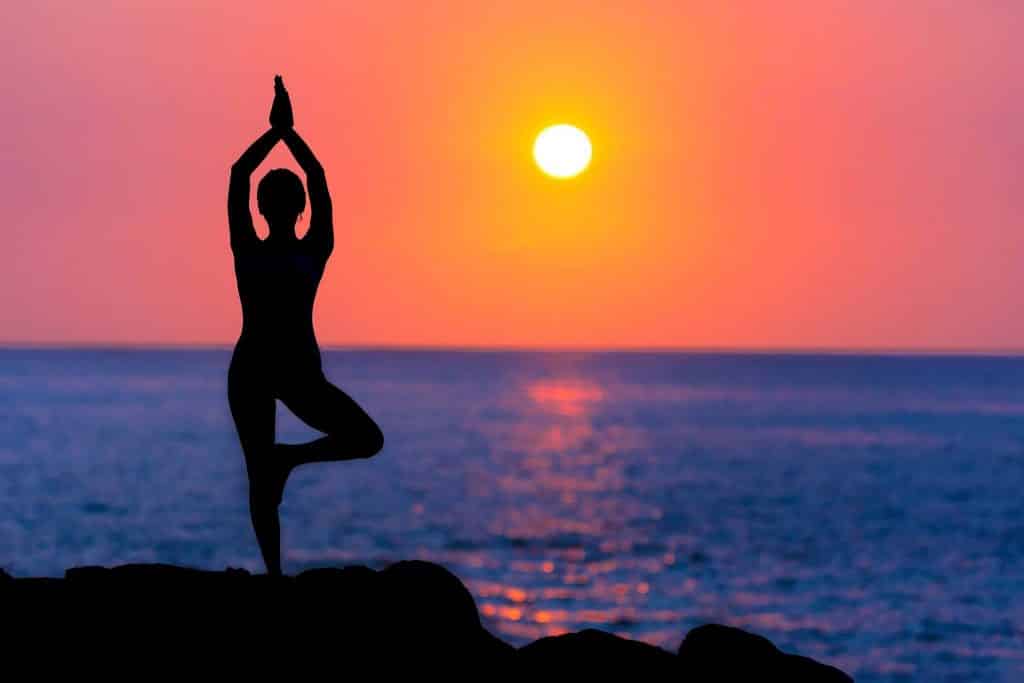 Silhueta de uma pessoa praticando a Vrksasana em frente ao mar, ao pôr do sol.