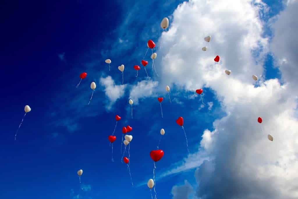 Imagem do céu azul e com poucas nuvens representando a reciprocidade. Vários balões em formato de coração nas cores vermelho e branco estão soltos no céu.
