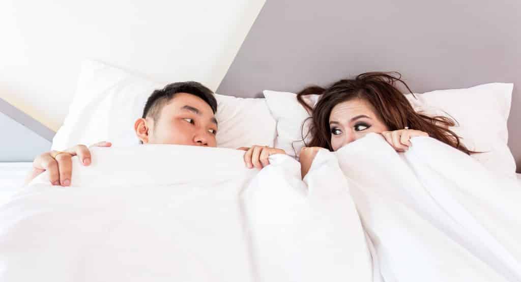 Imagem de um casal (homem e mulher) deitados em uma cama.  Um olhando para o outro.
