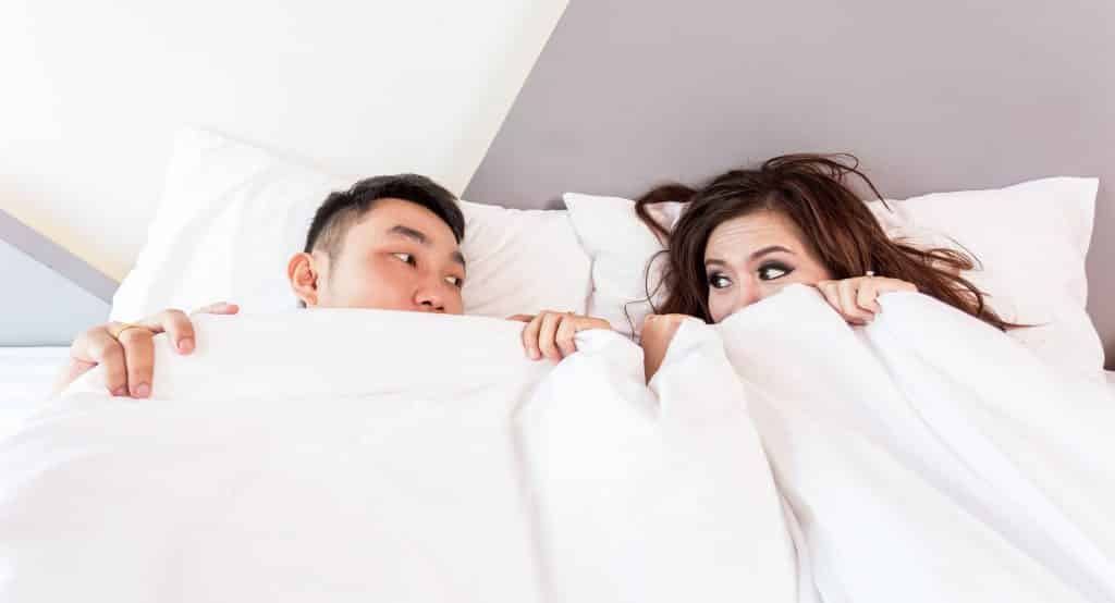 Imagem de um casal (homem e mulher) deitados em uma cama.  Um olhando para o outro.