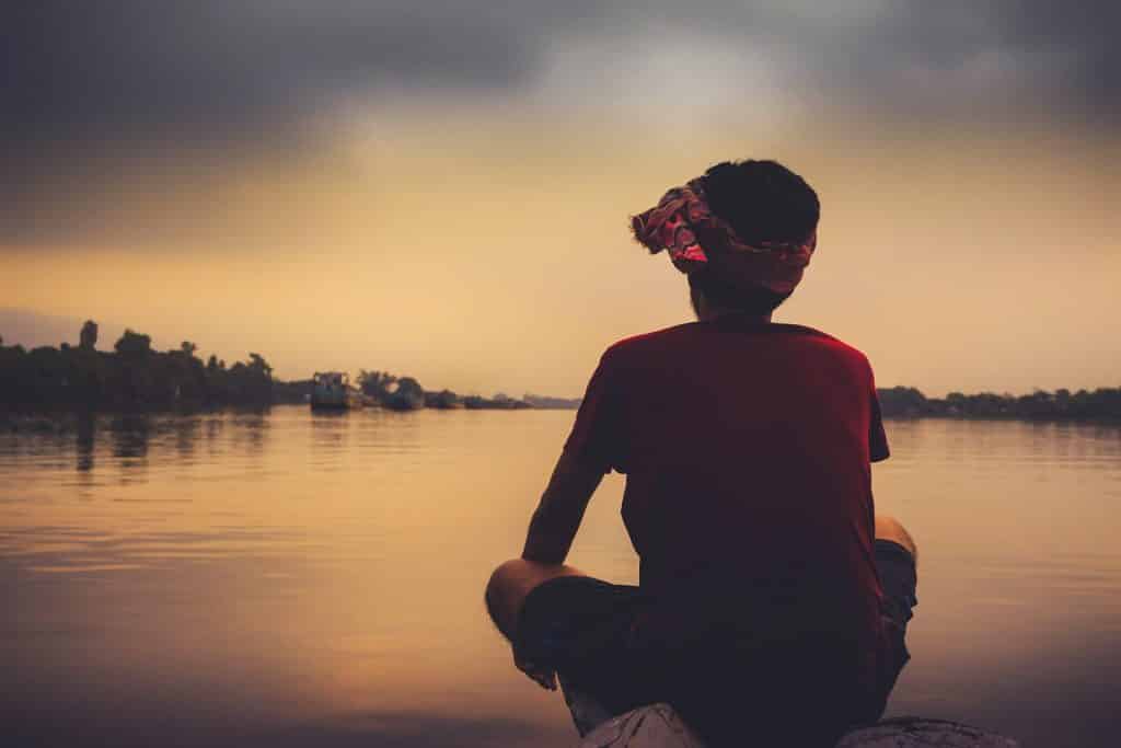 Imagem de um final de tarde em frente ao um grande rio. Na beira dele está um jovem garoto triste e solitário. Parece que está depressivo.