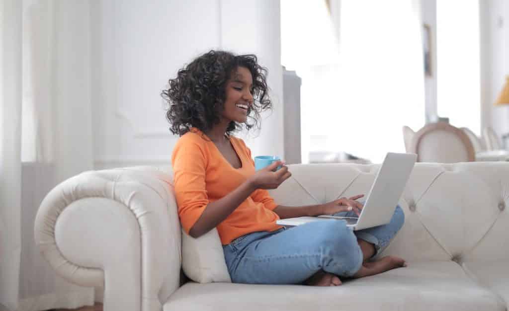 Mulher sentada em sofá, com notebook no colo, rindo enquanto digita.