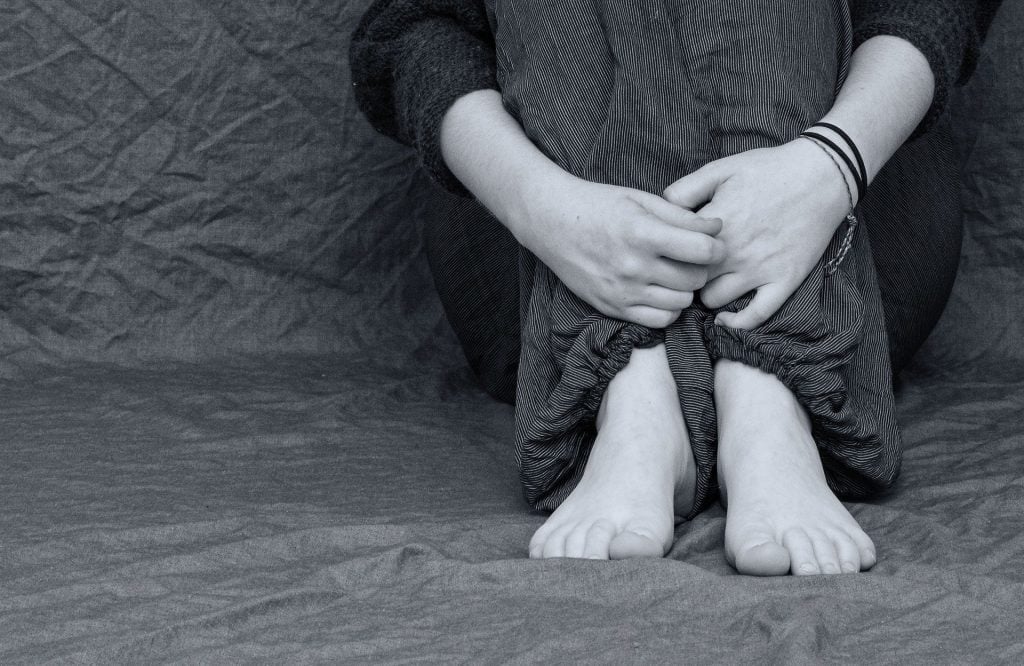 Imagem preto e brando de uma pessoa sentada e os braços abraçando as pernas. A pessoa está descalça e triste, deprimida.
