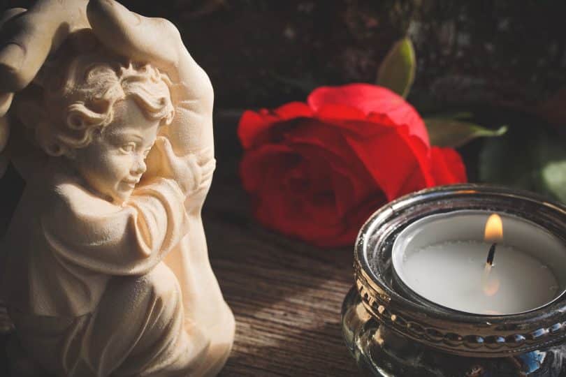 Imagem de um anjinho, uma rosa vermeha e uma vela, elementos que estão representando a religiosidade das pessoas.