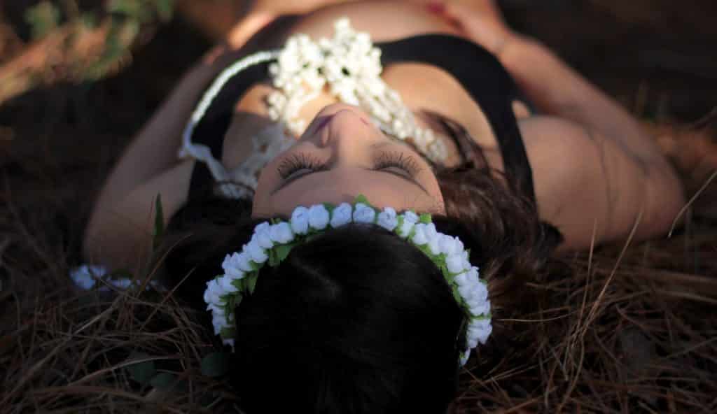 Imagem de uma mulher grávida usando um top preto e com uma tiara de flores na sua cabeça. Ela está deitada no gramado.