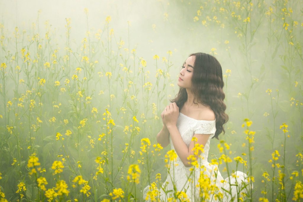 Imagem de um campo com flores amarelas em pleno nevoeira e uma mulher usando um vestido branco de renda fazendo a oração do perdão.
