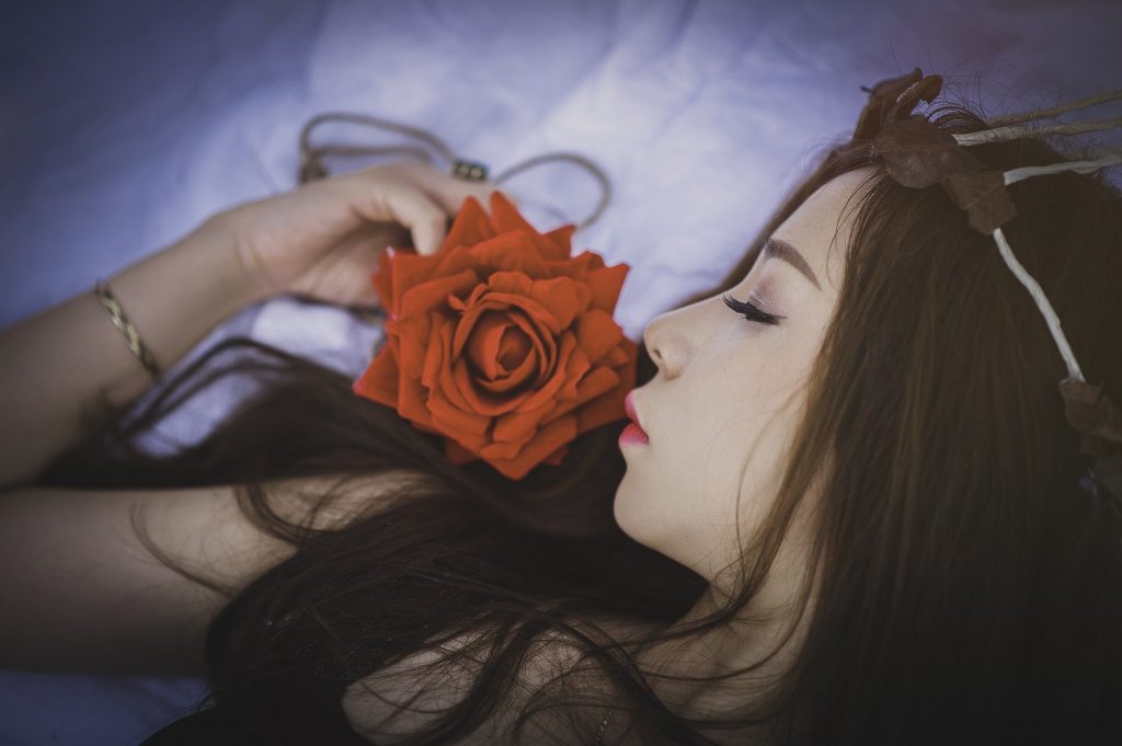 Imagem de uma linda jovem de cabelos longos deitada sobre a cama com lençol branco. Ao lado dela uma grande e maravilhosa rosa vermelha. A moça está com os olhos fechados. A imagem representa o orgasmo feminino.