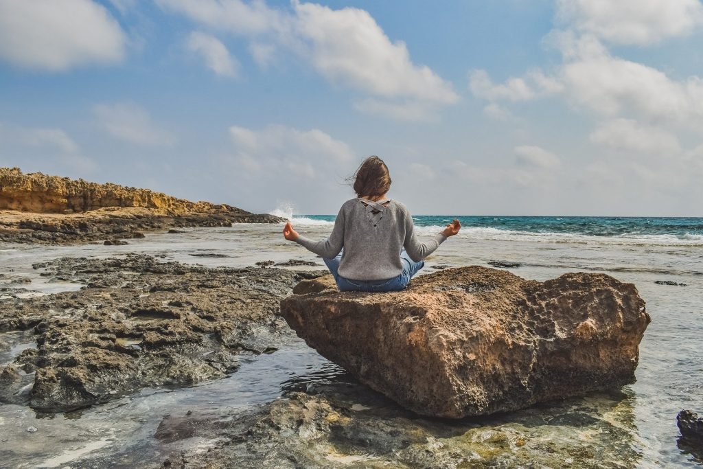 Imagem de uma mulher sentada de costas em uma pedra grande próximo ao mar. Ela está em posição de meditação.
