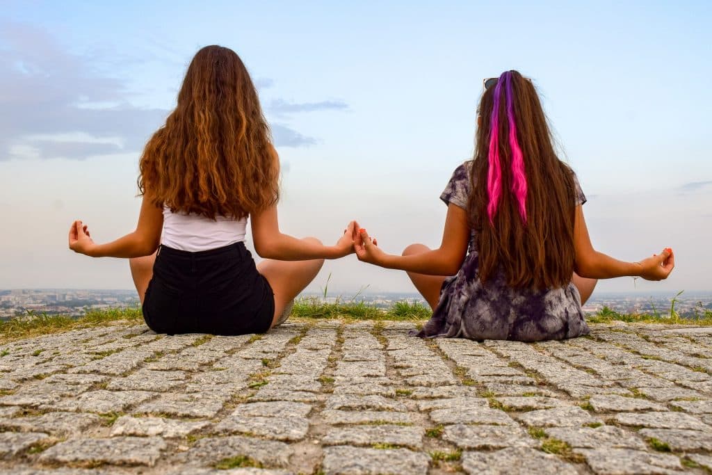 Imagem de duas garotas de costas sentadas no chão fazendo o uso da meditação.
