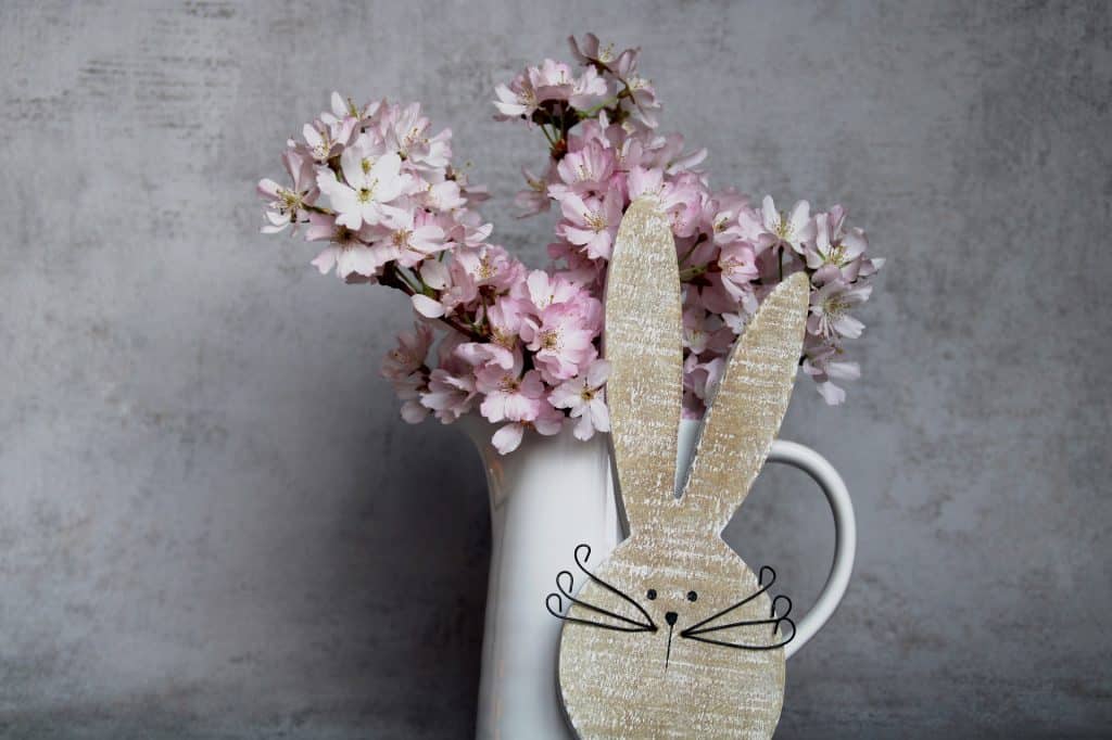 Imagem de um lindo vaso branco de porcelana cheio de flores na cor rosa. Ao lado a fprma da carinha do coelho da Páscoa.