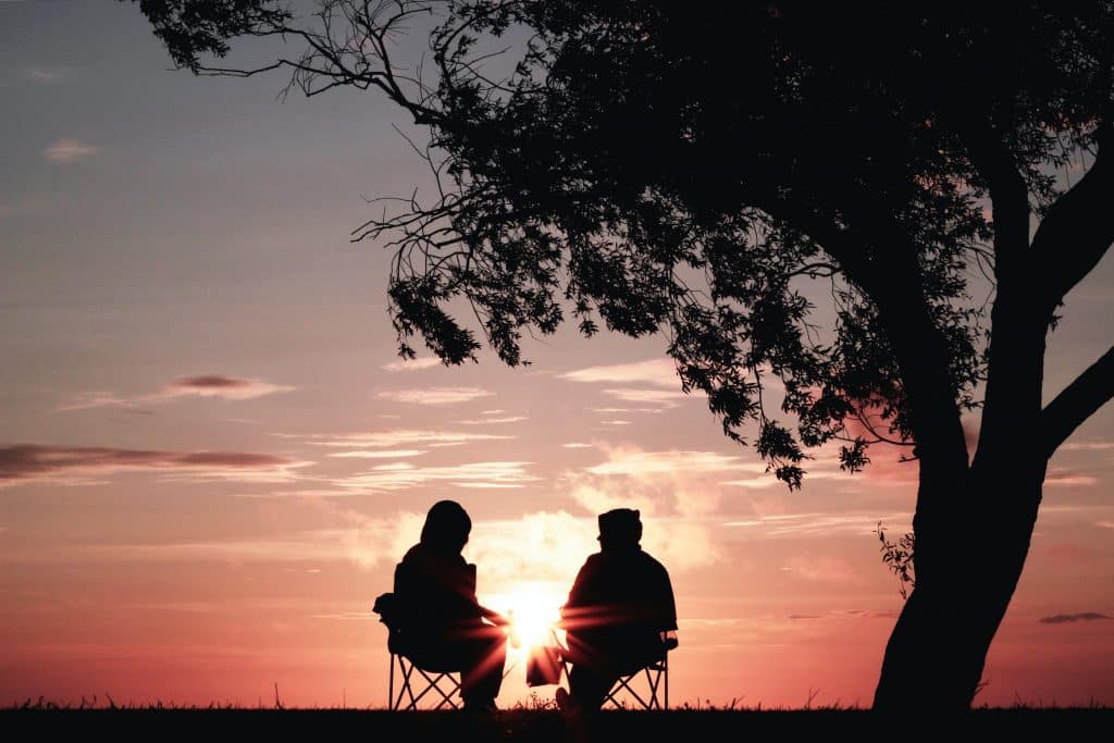 Duas pessoas conversando ao lado de uma árvore olhando o pôr do sol