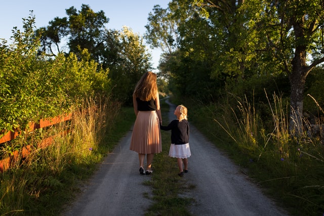 Mãe e filha de mãos dadas andando de costas em estrada no meio de árvores