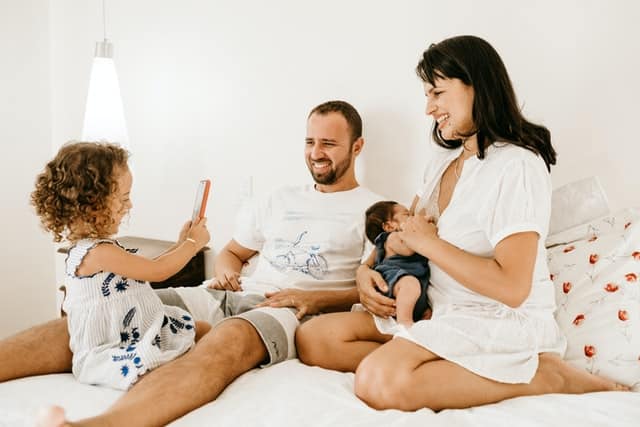 Família sentada na cama com pai, mãe, filha pequena e bebê mamando.