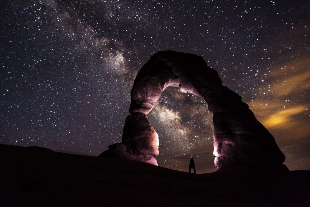 Homem em pé sob um arco de rocha, à noite, em meio a um céu estrelado.