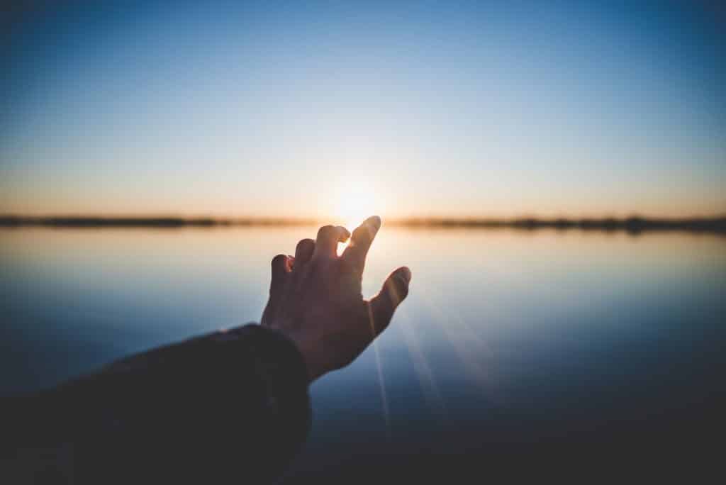 Pessoa com a mão esticada em direção ao sol e ao mar 