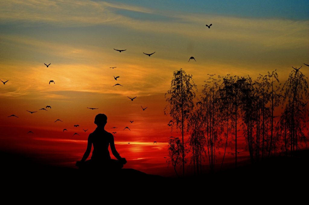 Imagem do por do sol e uma mulher sentada em uma pedra fazendo uma meditação. Ao fundo, vários pássaros sobrevoam o céu vermelho.