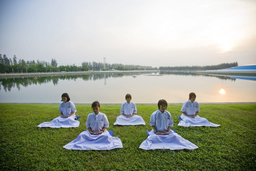 Imagem de cinco crianças vestidas com roupas brancas e sentadas em um gramado em posição de meditação.