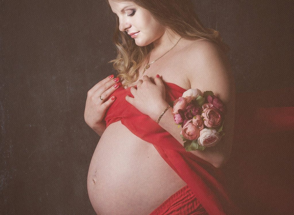 Imagem de uma linda mulher grávida e de cabelos longos. Ela está olhando para a sua barriga e em uma das mãos ela segura m lindo buquê de rosas.
