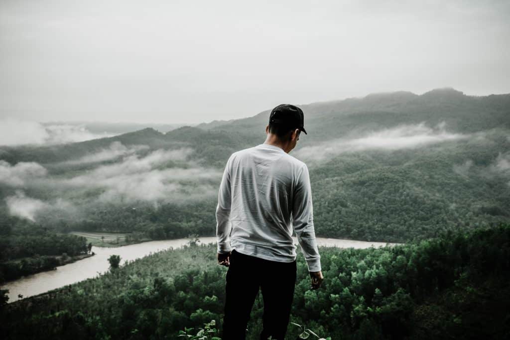 Homem visto de costas em pé em um lugar alto, com uma paisagem natural a sua volta.