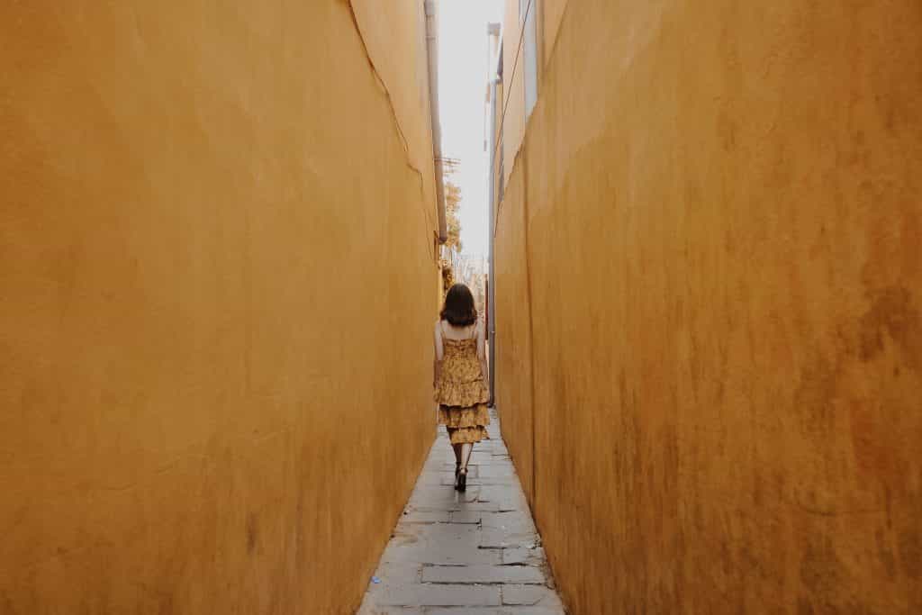 Mulher de vestido caminhando por um corredor estreito, vista de costas.