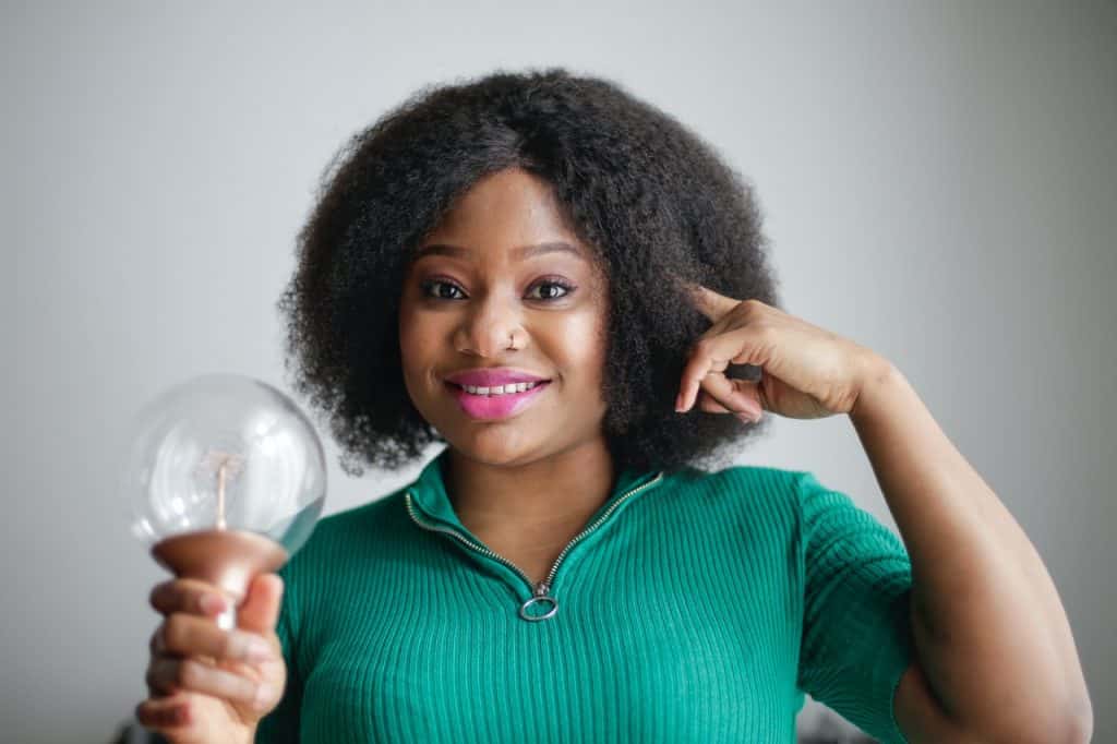 Mulher sorrindo, com um dedo indicador ao lado da cabeça, e segurando uma lâmpada, indicando uma ideia.