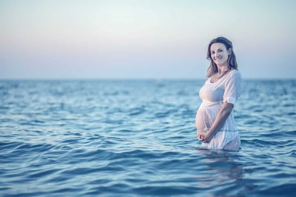 Imagem de um lindo mar azul e dentro dele está uma mulher grávida vestindo um vestido branco. Ela está segurando a sua barriga.
