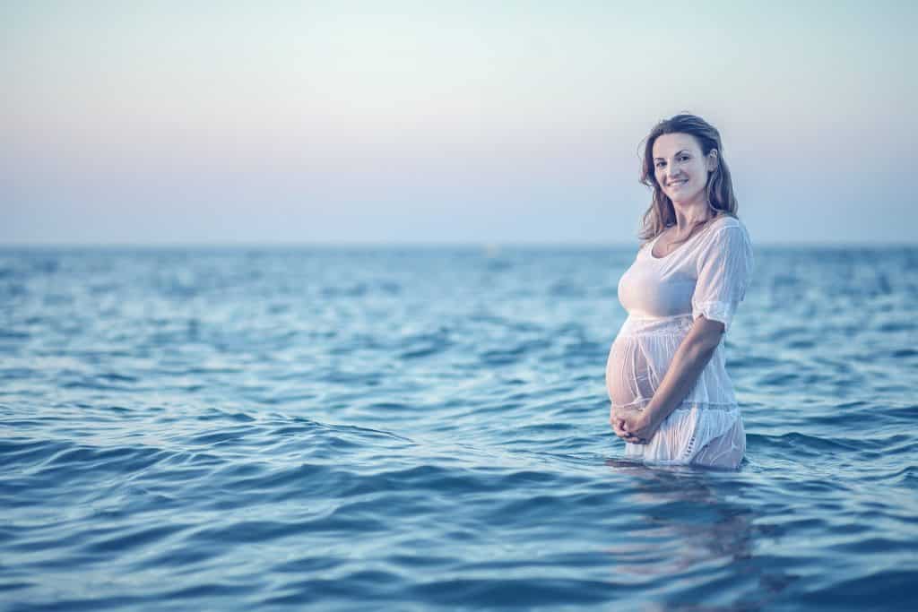 Imagem de um lindo mar azul e dentro dele está uma mulher grávida vestindo um vestido branco. Ela está segurando a sua barriga.