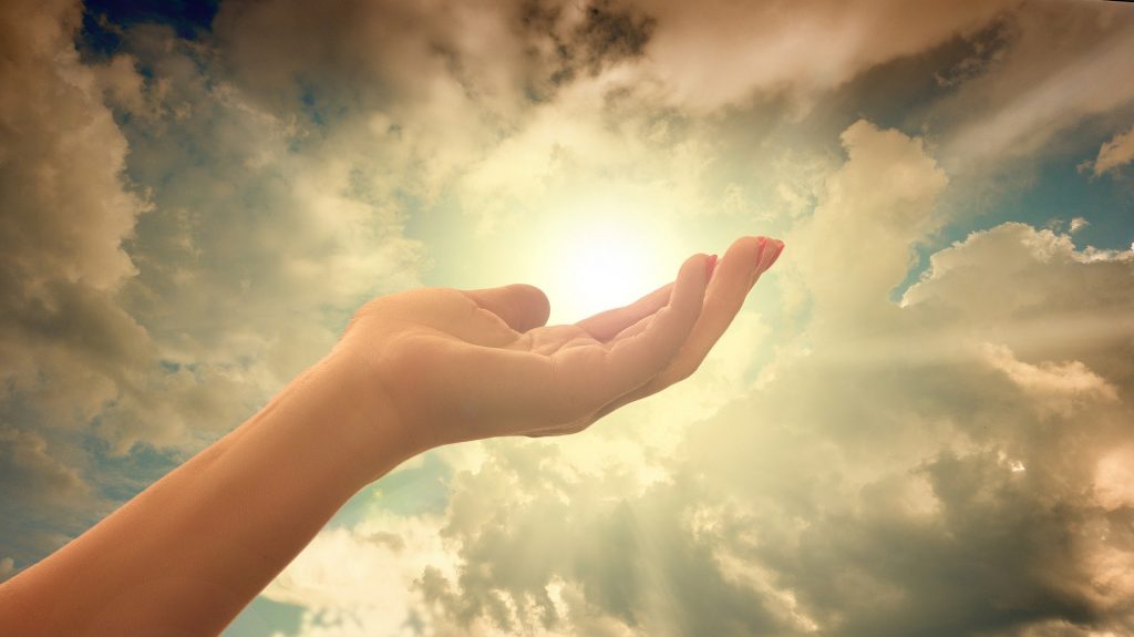 Imagem do céu com a luz do sol e muitas nuvens. Ao fundo a mão de uma mulher representando a sua espiritualidade.
