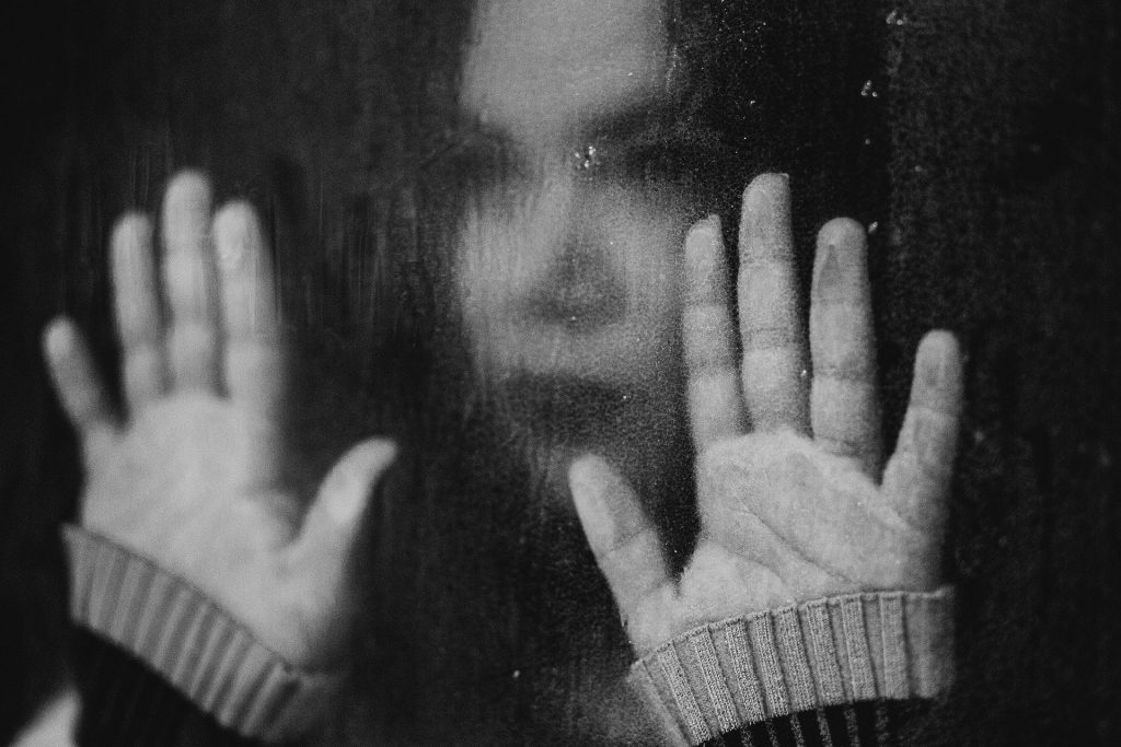 Imagem em preto e branco do rosto de uma mulher ansiosa refletida no vidro de uma janela. As mãos da mulher estão sobre o vidro.