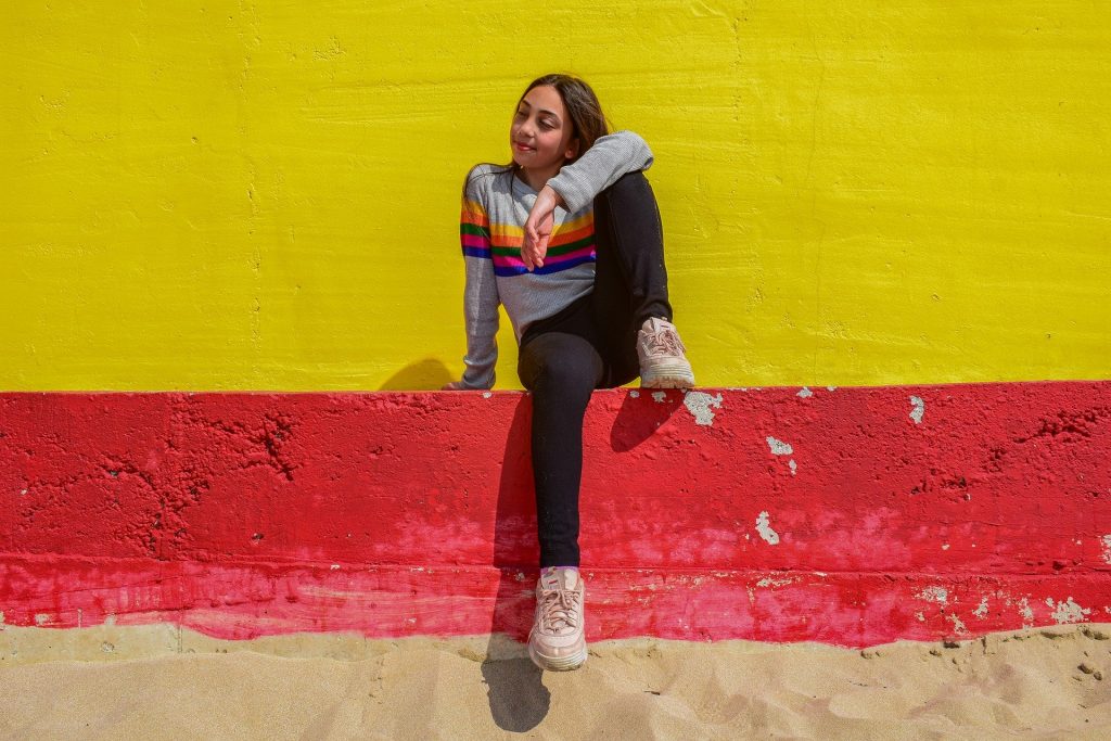 Imagem com fundo de uma parede pintada nas cores amarela e vermelha. À frente uma jovem, bem despreocupada, sentada e alegre.
