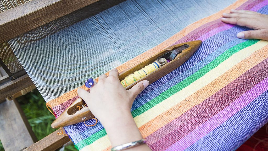 Imagem de das mãos de uma mulher em um tear tecendo um lindo trabalho colorido.
