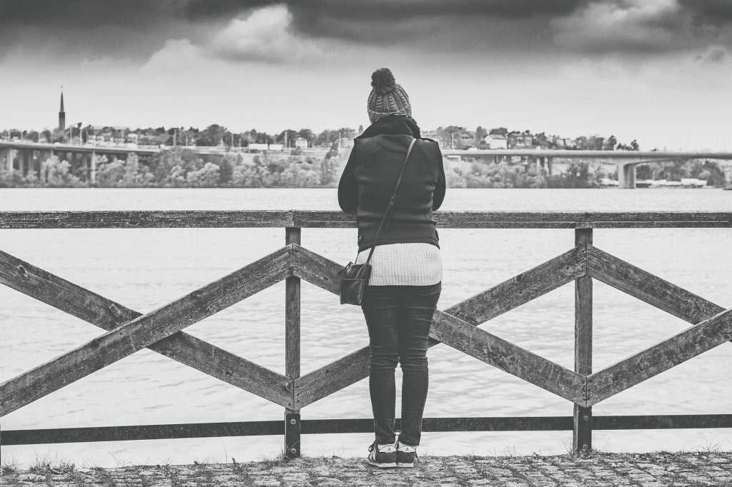 Imagem em preto e branco de um rio. Sobre ele uma ponte onde está uma mulher ansiosa. Ela usa roupas de inverno e uma touca e está olhando para o rio.
