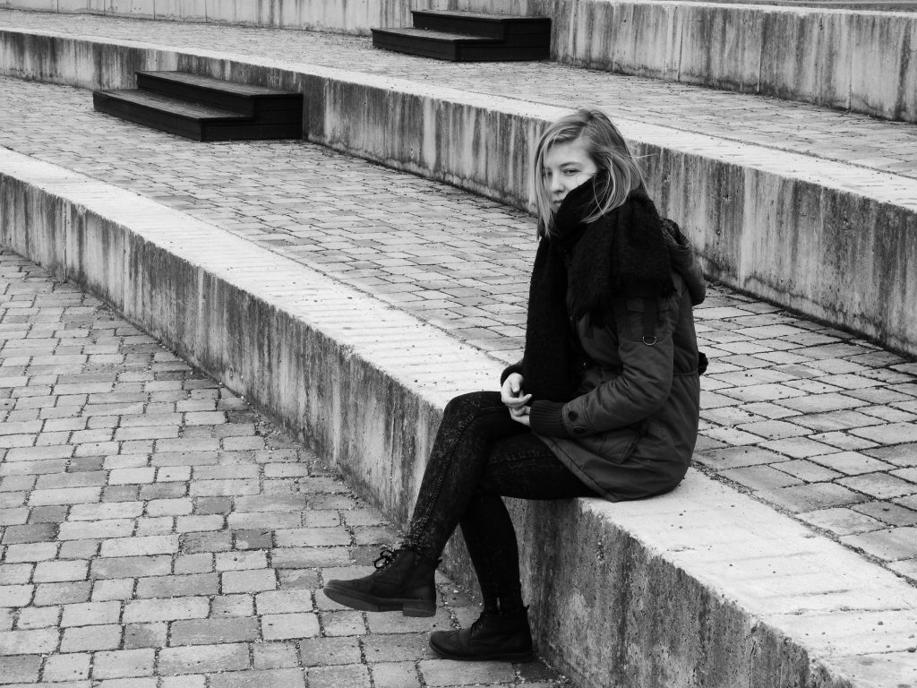 Imagem preto e branco de uma escadaria e uma mulher vestindo um look de inverno. Ela está sentada em um dos degraus da escadaria e parece estar muito triste e deprimida.
