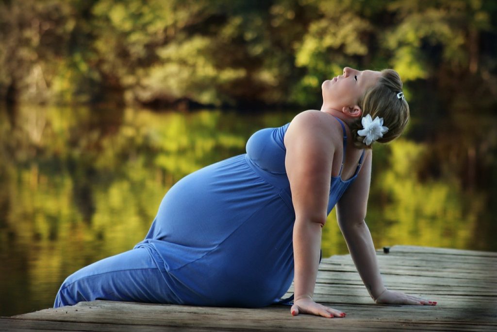 Imagem de uma mulher grávida com uma flor branca no seu cabelo, usando um lindo vestido azul. Ela está sentada no pier em frente ao lago.
