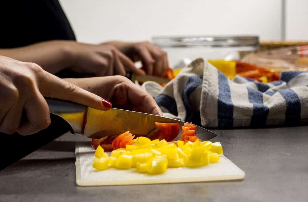 Imagem das mãos de uma mulher na cozinha. Ela está picando legumes sobre uma tábua branca.
