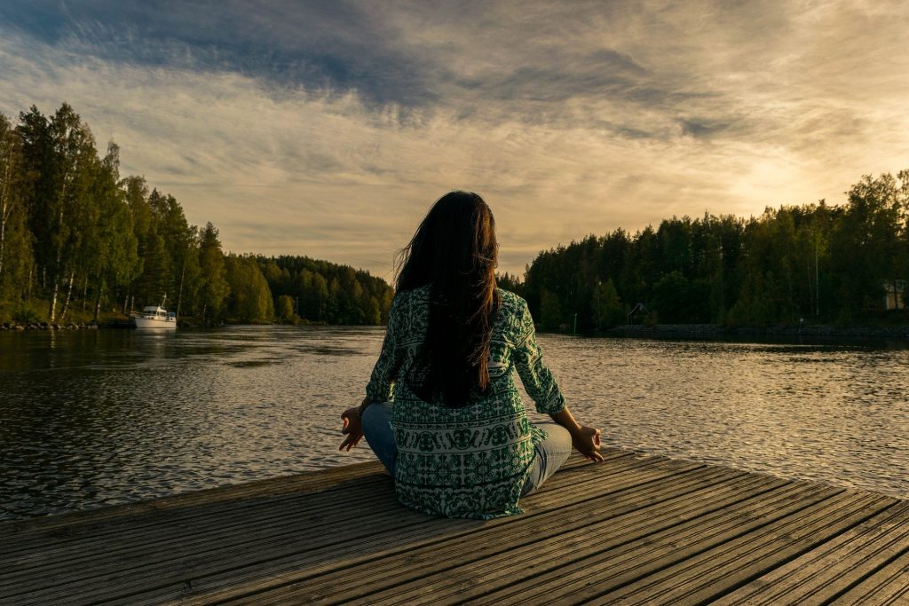 Imagem de uma mulher sentada em um pier de frente para um lago. Ela está sentada de costas em posição d meditação.