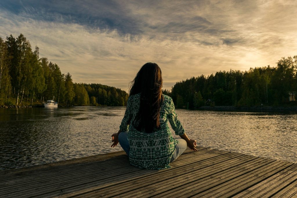 Imagem de uma mulher sentada em um pier de frente para um lago. Ela está sentada de costas em posição d meditação.
