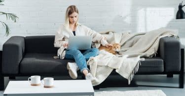Mulher no sofá com notebook no colo fazendo carinho em cachorro ao lado