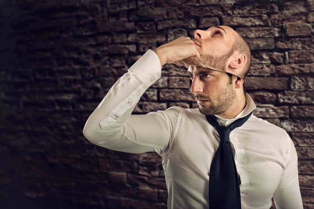 Homem vestido roupas sociais tira de seu rosto uma máscara com sua própria face.