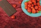 Imagem de um prato com lindos morangos vermelhos e uma barra de chocolate sobre um lindo tecido na cor vermelho. Os alimentos ajudam aumentar a libido.