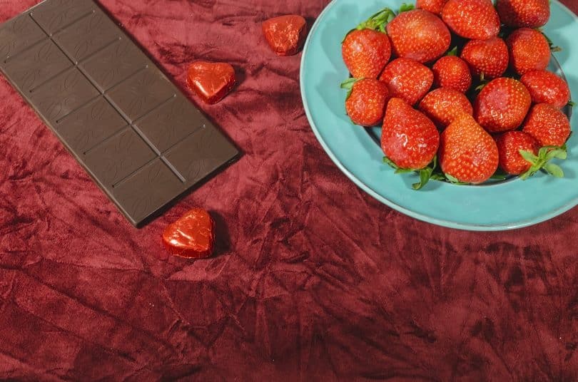 Imagem de um prato com lindos morangos vermelhos e uma barra de chocolate sobre um lindo tecido na cor vermelho. Os alimentos ajudam aumentar a libido.