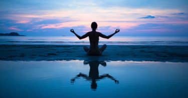 Mulher sentada meditando na praia com céu rosa e azul ao fundo