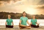 Três pessoas sentadas meditando em frente a um rio.