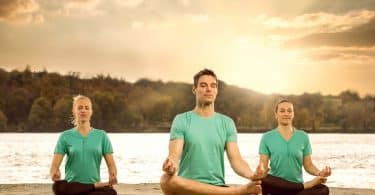 Três pessoas sentadas meditando em frente a um rio.