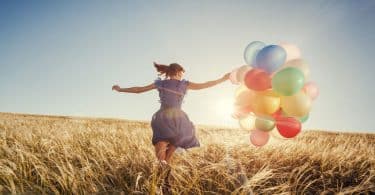 Menina correndo com balões coloridos em campo com sol refletindo