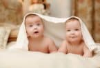 Imagem de dois bebês deitados de bruço com um lençol sobre a cabeça deles. Eles estão sorrindo.