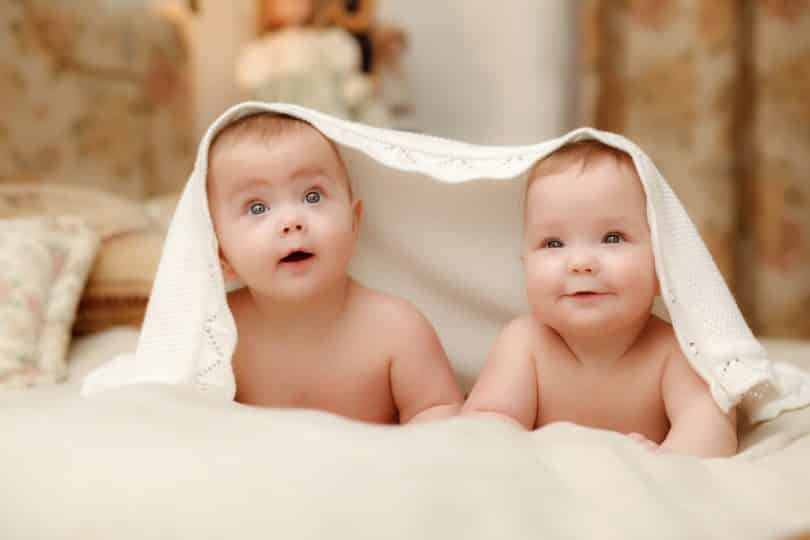 Imagem de dois bebês deitados de bruço com um lençol sobre a cabeça deles. Eles estão sorrindo.