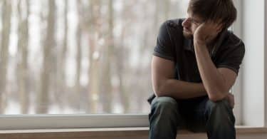 Homem sentado ao lado de uma janela apoiando sua cabeça com seu braço com rosto de tristeza