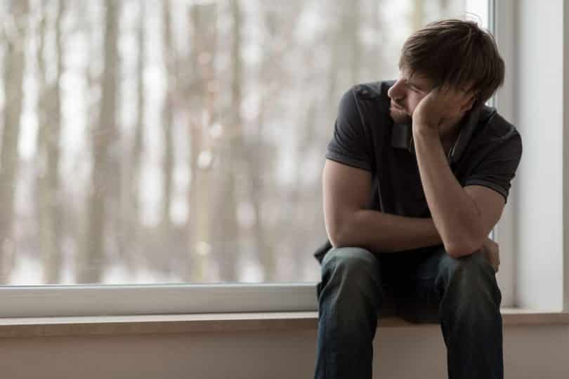 Homem sentado ao lado de uma janela apoiando sua cabeça com seu braço com rosto de tristeza