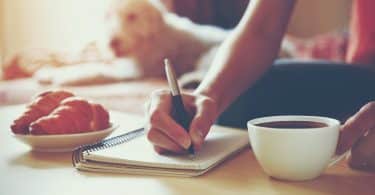 Mulher segurando uma caneta e escrevendo em seu caderno ao lado de uma xícara de café